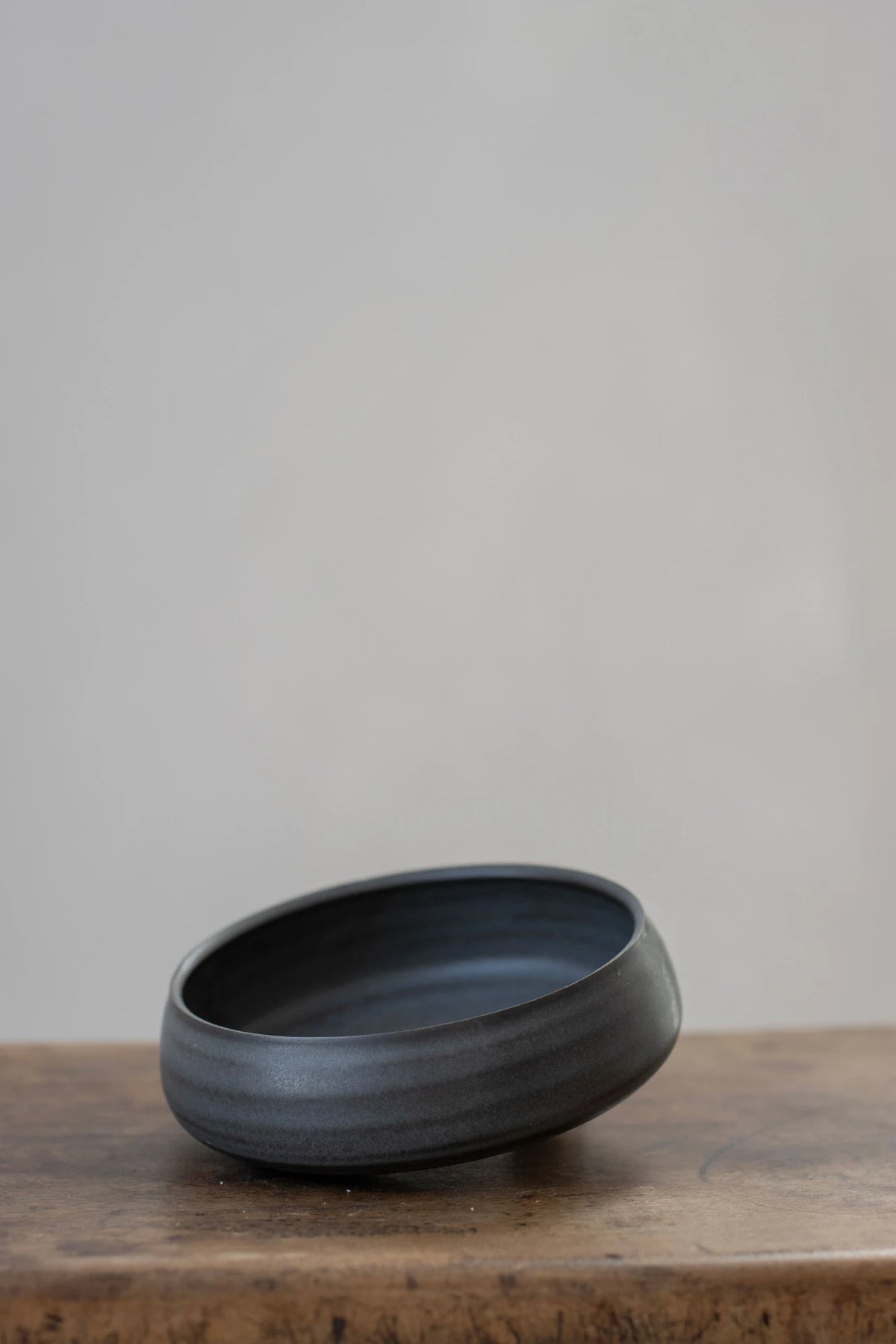 A Borja Moronta Curved side bowl in nightshade blue glaze, resting on a rustic wooden surface against a neutral light grey background.