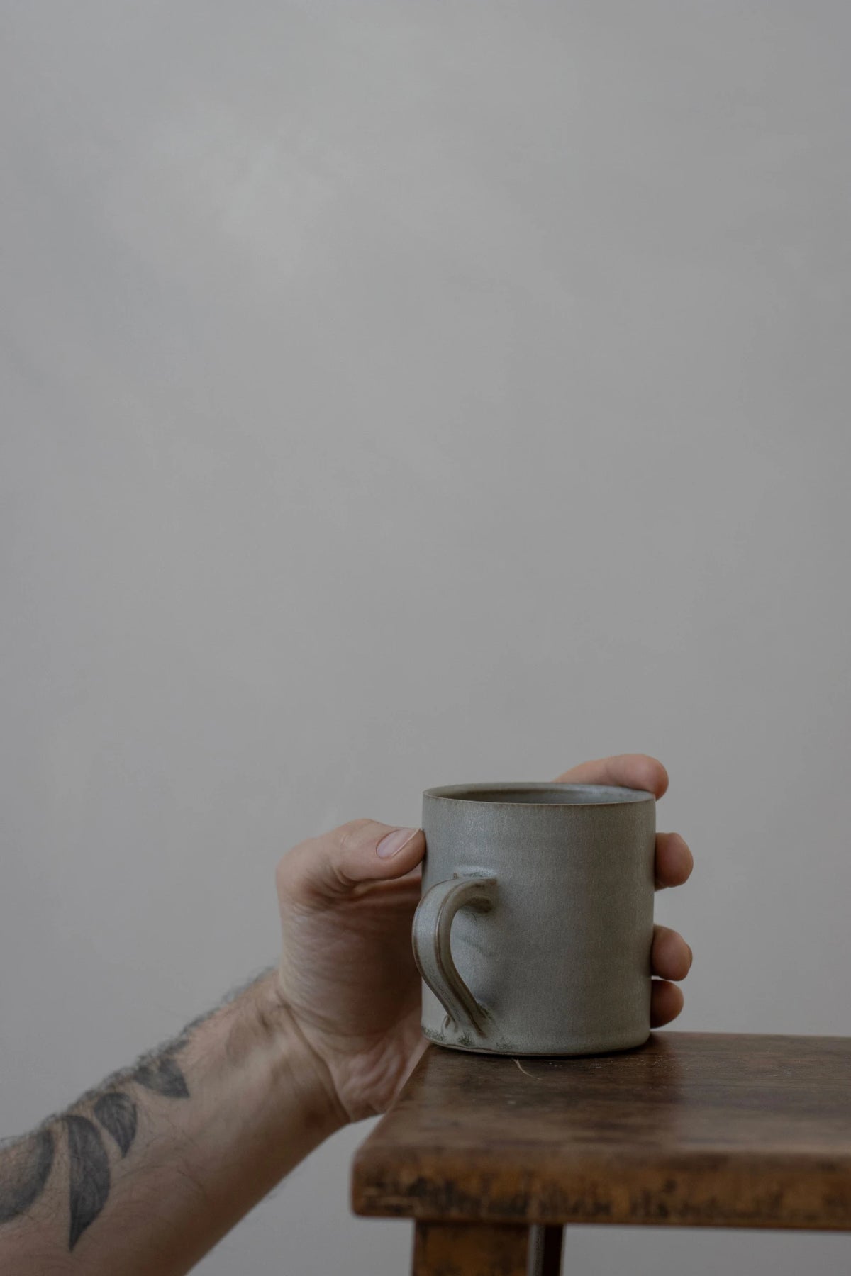 A hand with a tattoo holds a Borja Moronta cylindrical cup in feather grey glaze resting on a wooden table against a plain grey wall.