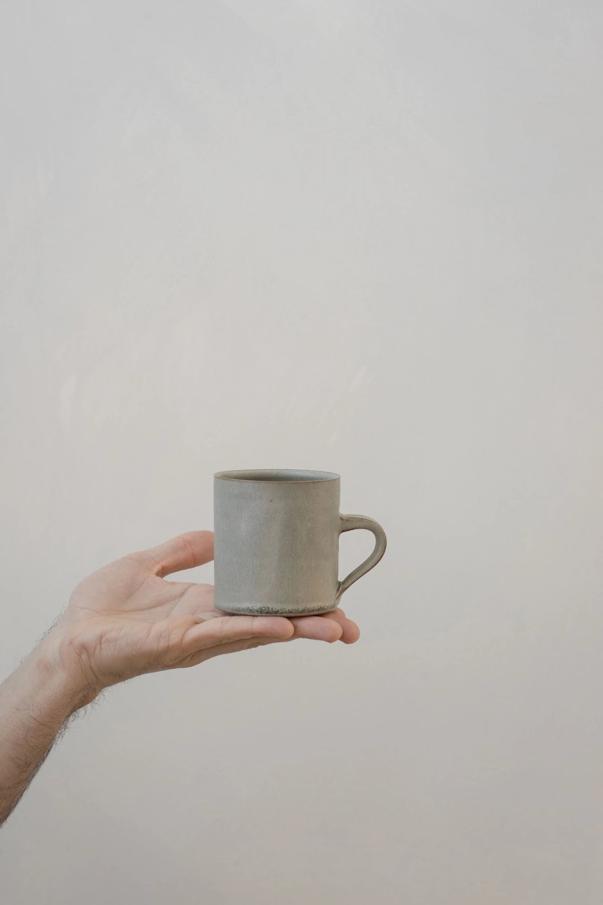 A hand holding a Borja Moronta cylindrical cup in Feather Grey glaze against a plain, light beige background.