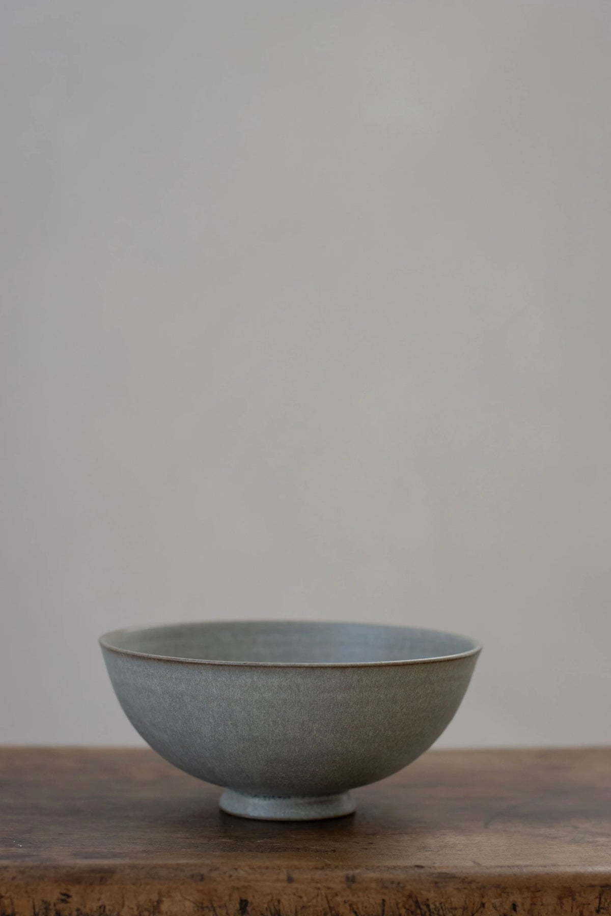 A Borja Moronta small footed bowl in Mist Blue glaze, sitting on a rustic wooden surface against a neutral, warm grey background.