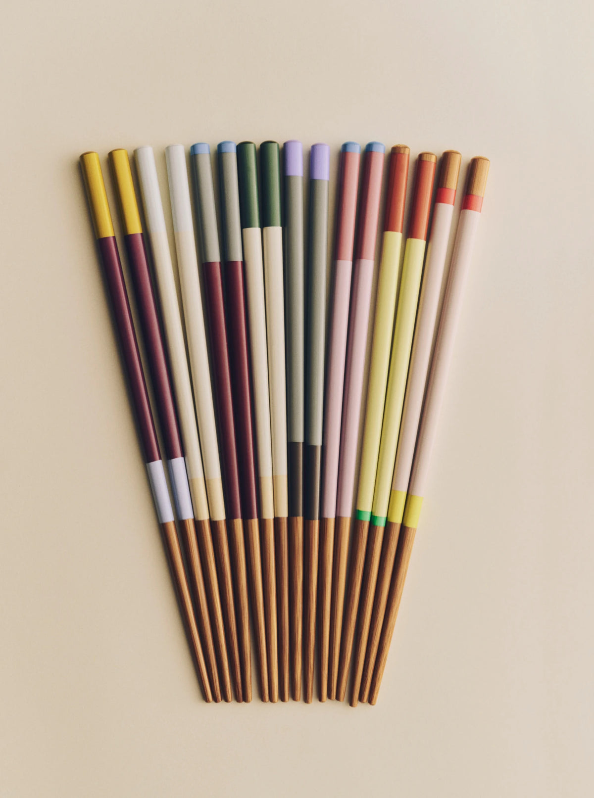 A fan arrangement of colorful chopsticks on a warm beige background. The chopsticks feature various color-blocked sections in yellow, white, green, purple, red, pink, and brown wood at the tips and handles.