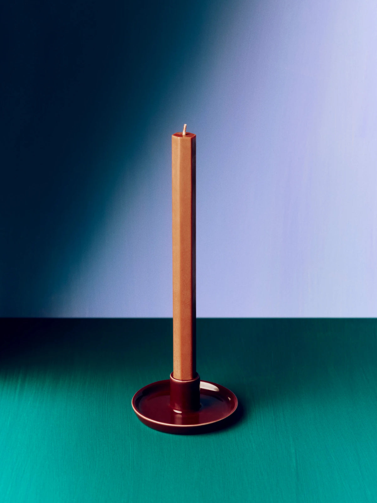 A tall, slender brown taper candle in a matching chestnut brown HAY disc candle holder, set against a split blue and teal background.