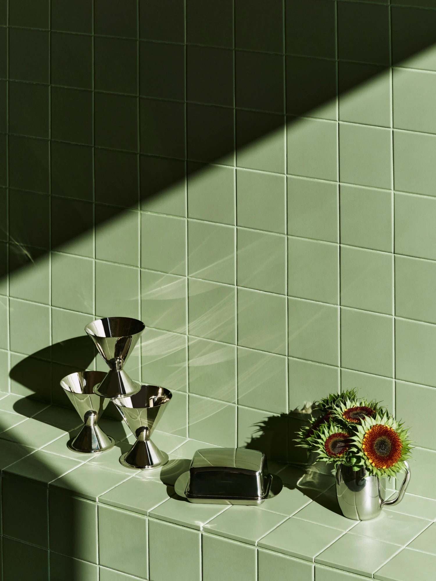 HAY stainless steel ice cream cups, butter box and jug with sunflowers on a green tiled wall with sunlight casting shadows.