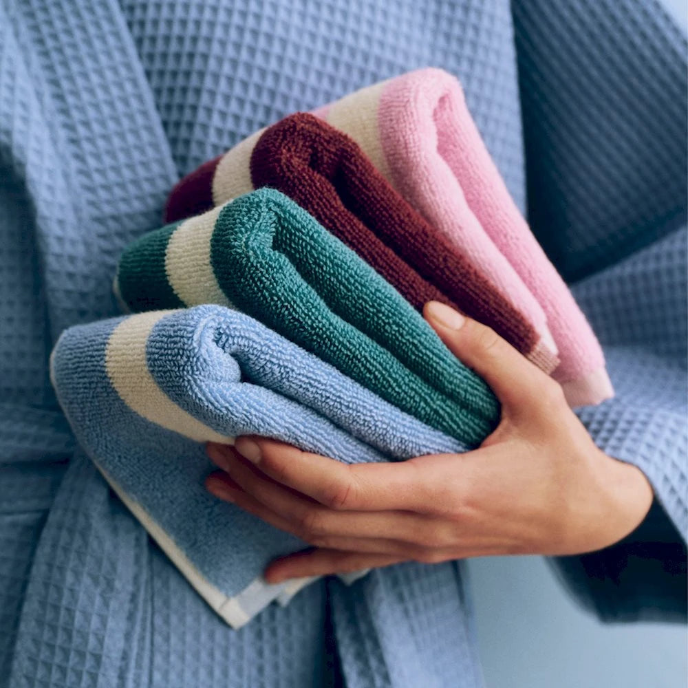A hand holding 3 colours of folded HAY stripe cotton wash cloths against a blue waffle bathrobe