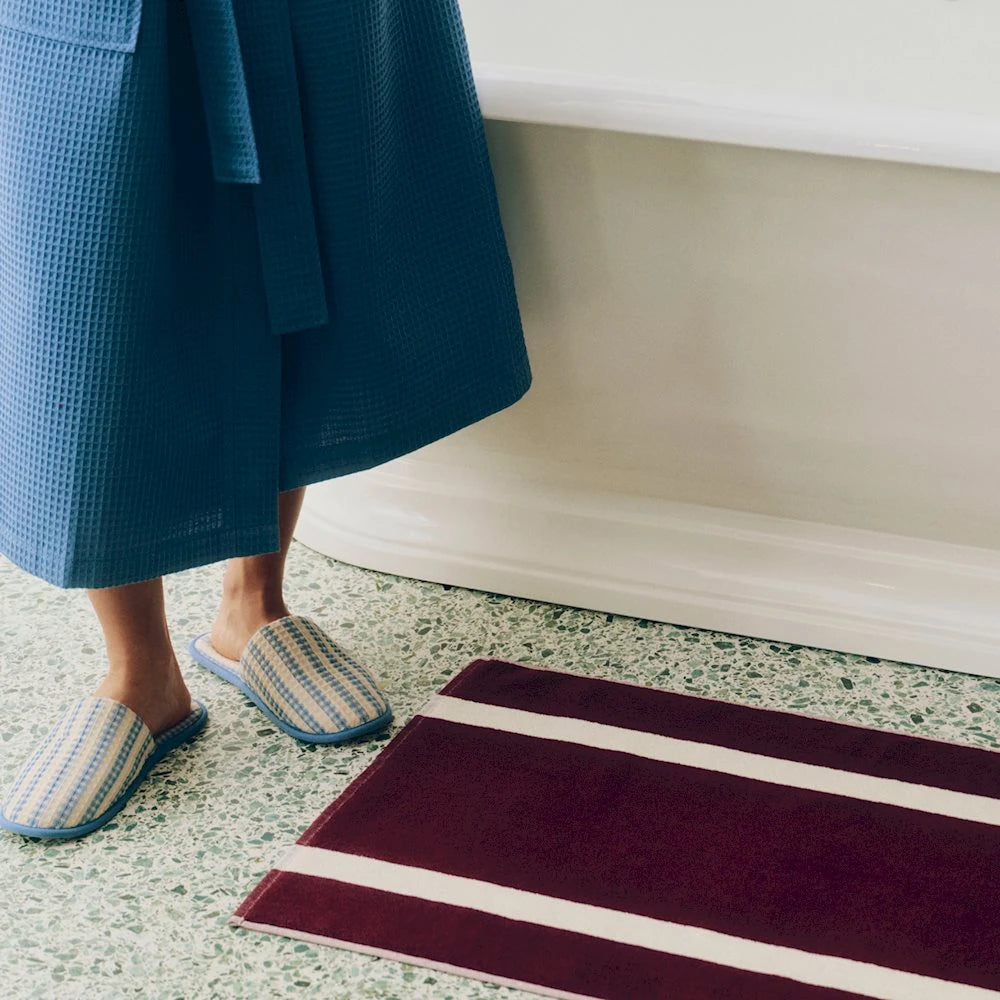 A person wearing a HAY blue waffle-knit robe and matching dusty blue HAY waffle slippers stands on a speckled bathroom floor next to a bathtub, with a burgundy bath mat featuring white stripes in the foreground.