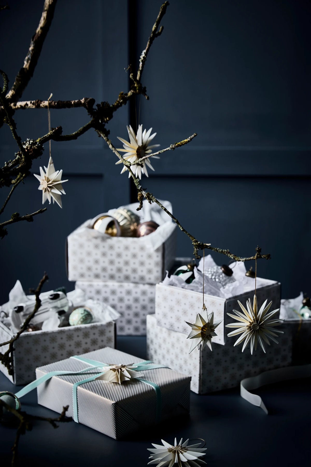 A styled arrangement of wrapped gift boxes and open boxes with decorative white patterned paper, metallic ornaments, and hanging star-like ornaments on dark branches against a dark blue background.