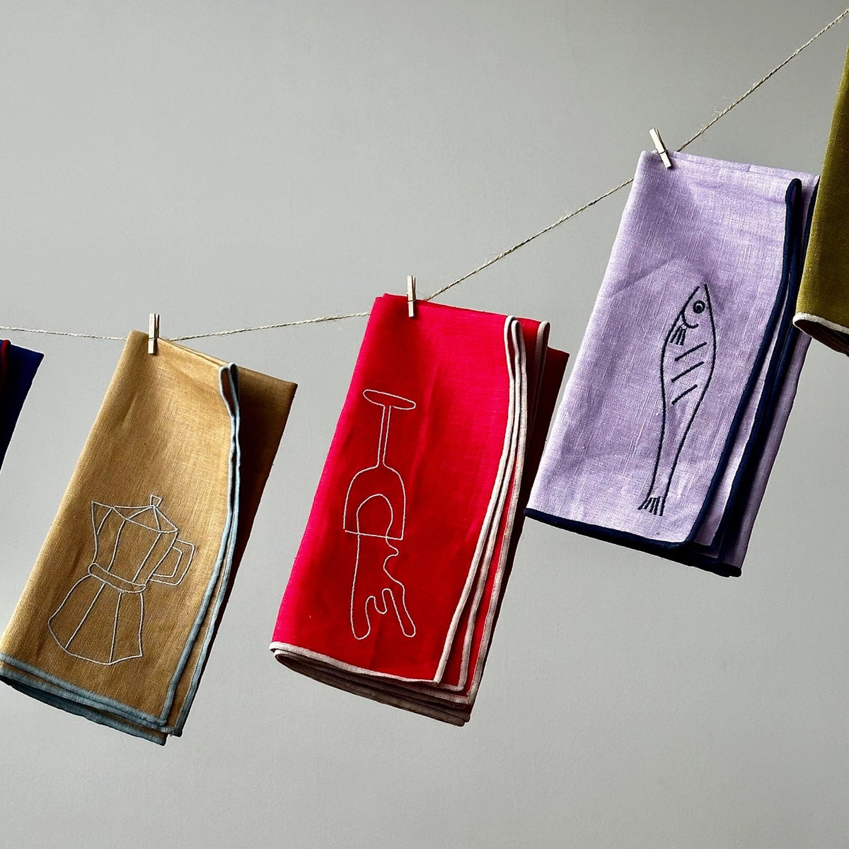 Colorful embroidered napkins by Maeganhanging on a line against a gray background
