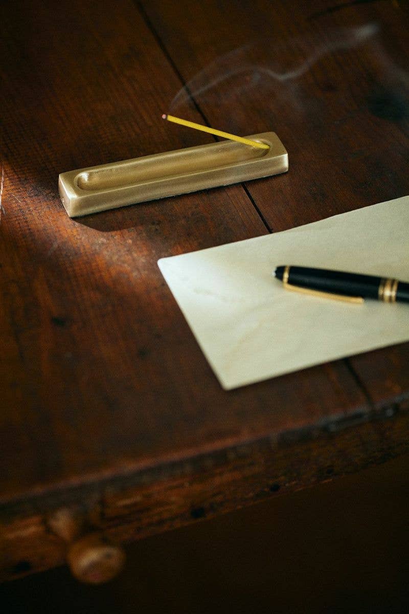 Incense stick burning in a brass holder on a wooden surface with a pen and paper.