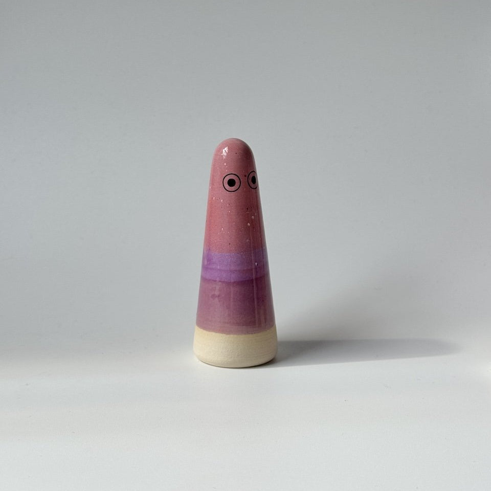 Ely Studio Arhoj ghost, featuring a pink and purple glazed body with a natural beige wooden base and simple painted eyes, set against a plain light grey background.