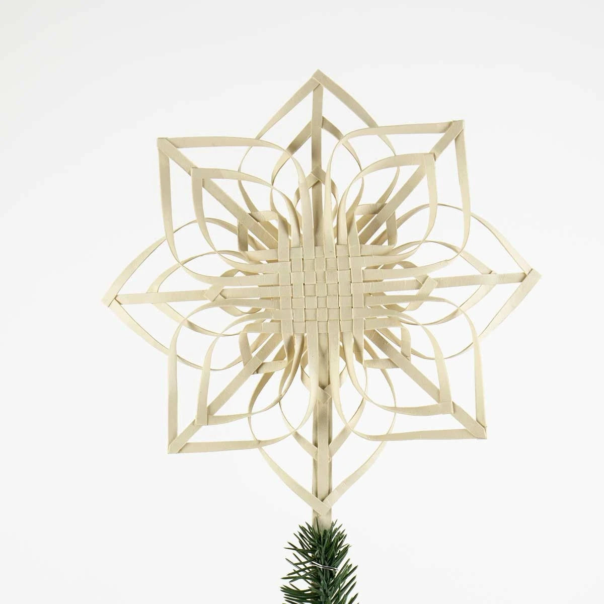 A light natural off white paper Christmas tree topper shaped like a multi-pointed geometric star, shown with a small section of green pine branch at the bottom against a plain white background.