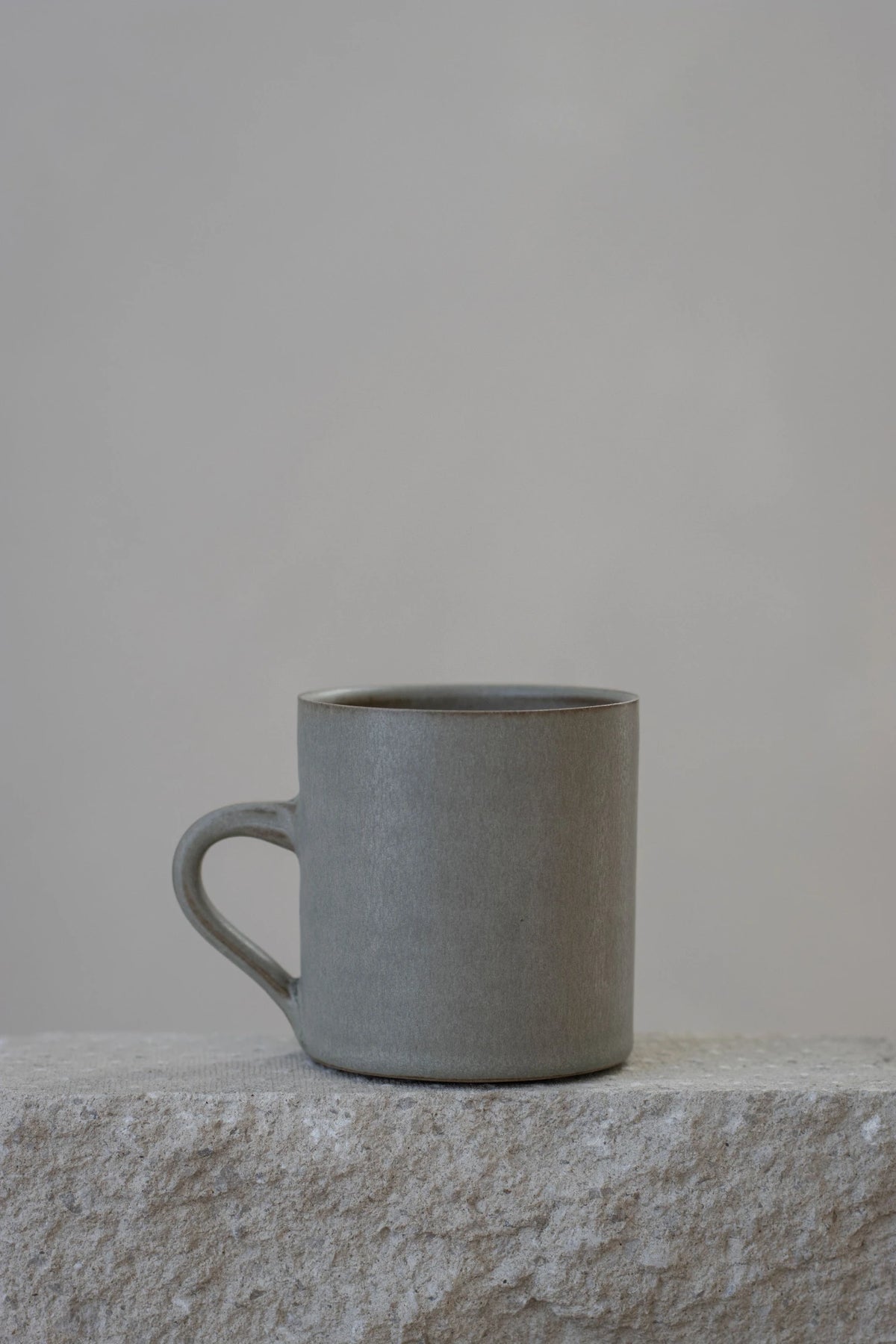 A Borja Moronta Cylindrical cup in Feather Grey Glaze, resting on a rough stone ledge against a neutral light gray background.