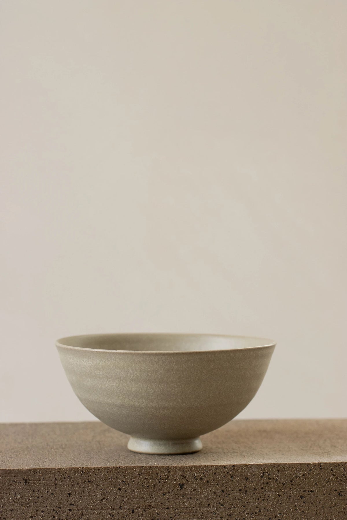 A Borja Moronta footed bowl in Pearl Grey glaze, resting on a textured concrete surface against a solid, warm off-white background.