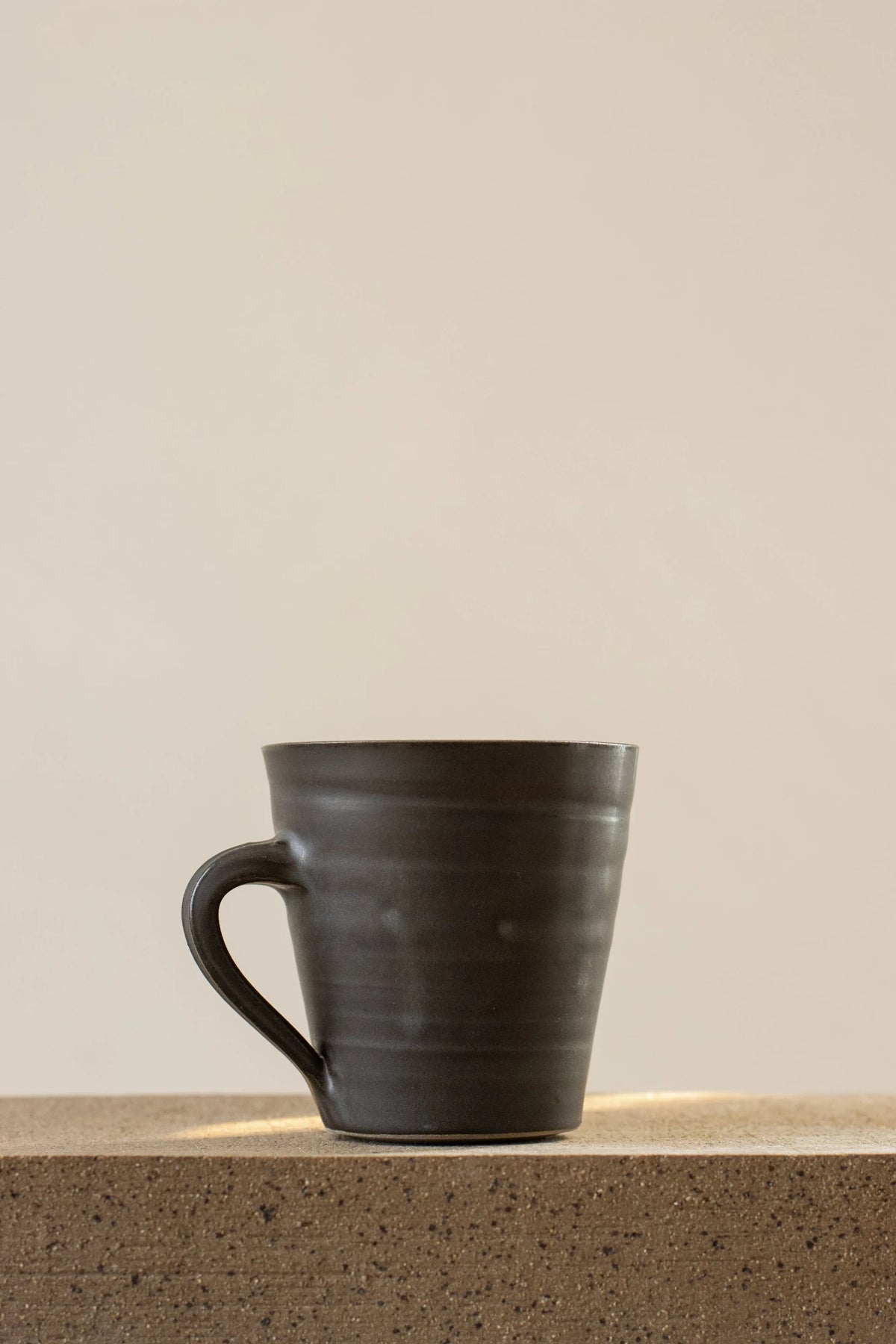 A Borja Moronta flared mug in nightshade blie glaze, resting on a textured concrete surface against a warm beige background.