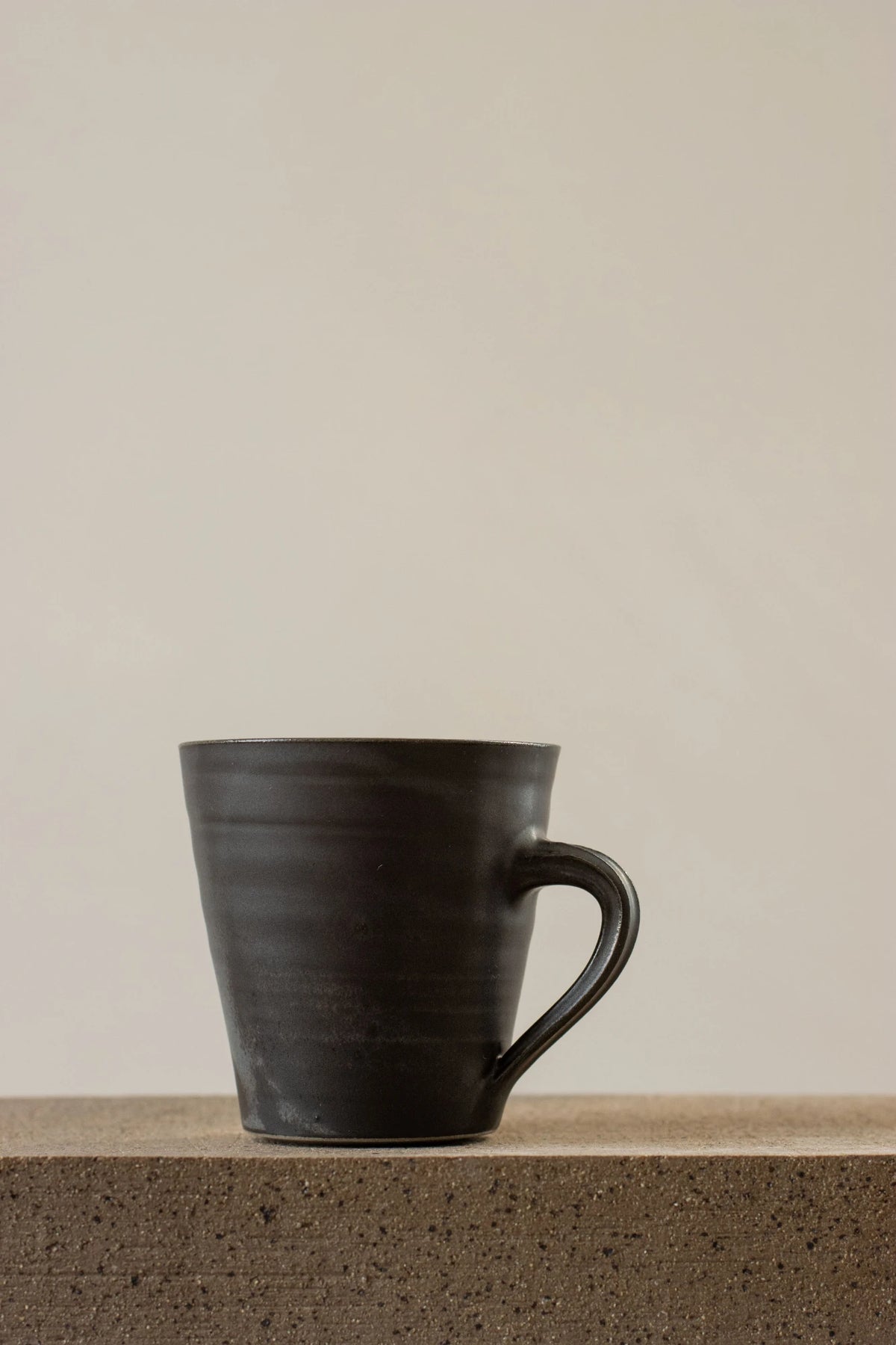 A Borja Moronta flared mug in Nightshade Blue glaze, resting on a textured concrete ledge against a solid beige background.