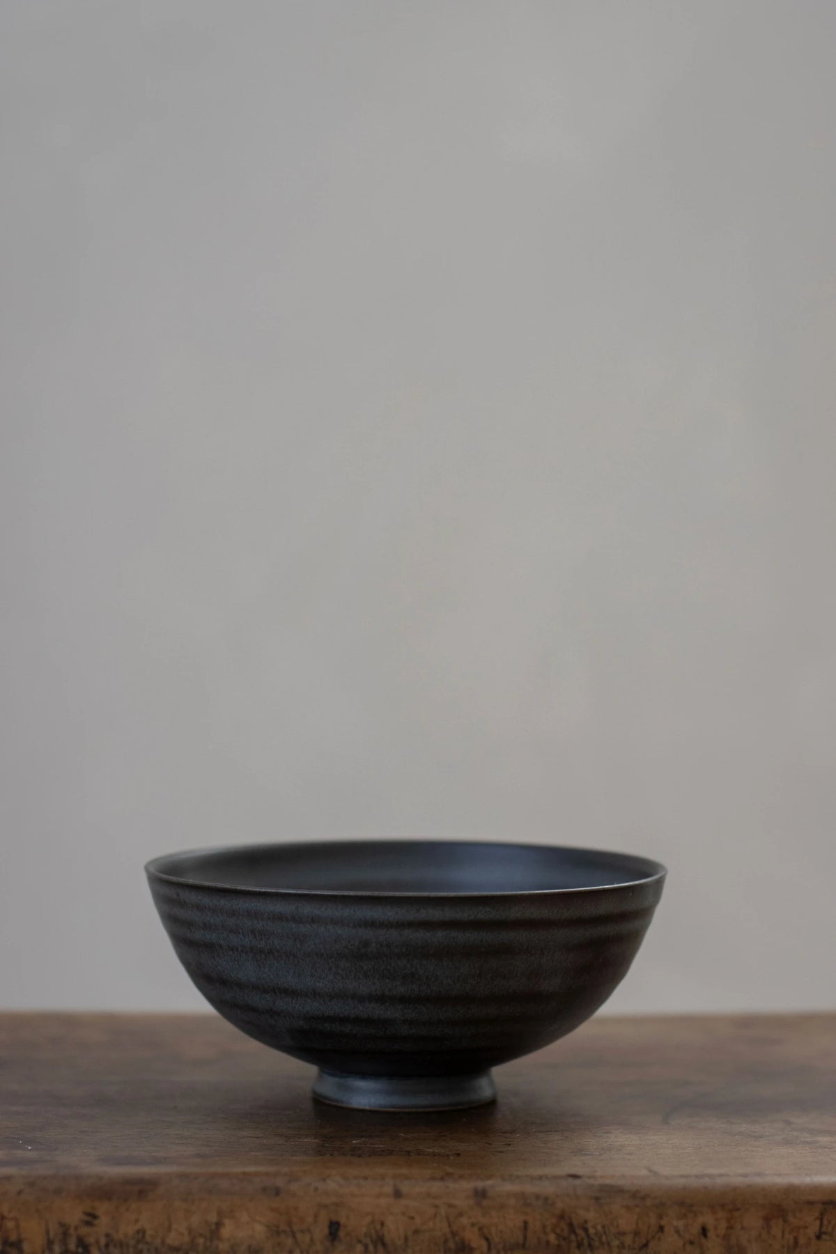 A Borja Moronta small footed bowl in nightshade blue, resting on a rustic wooden surface against a neutral gray background.