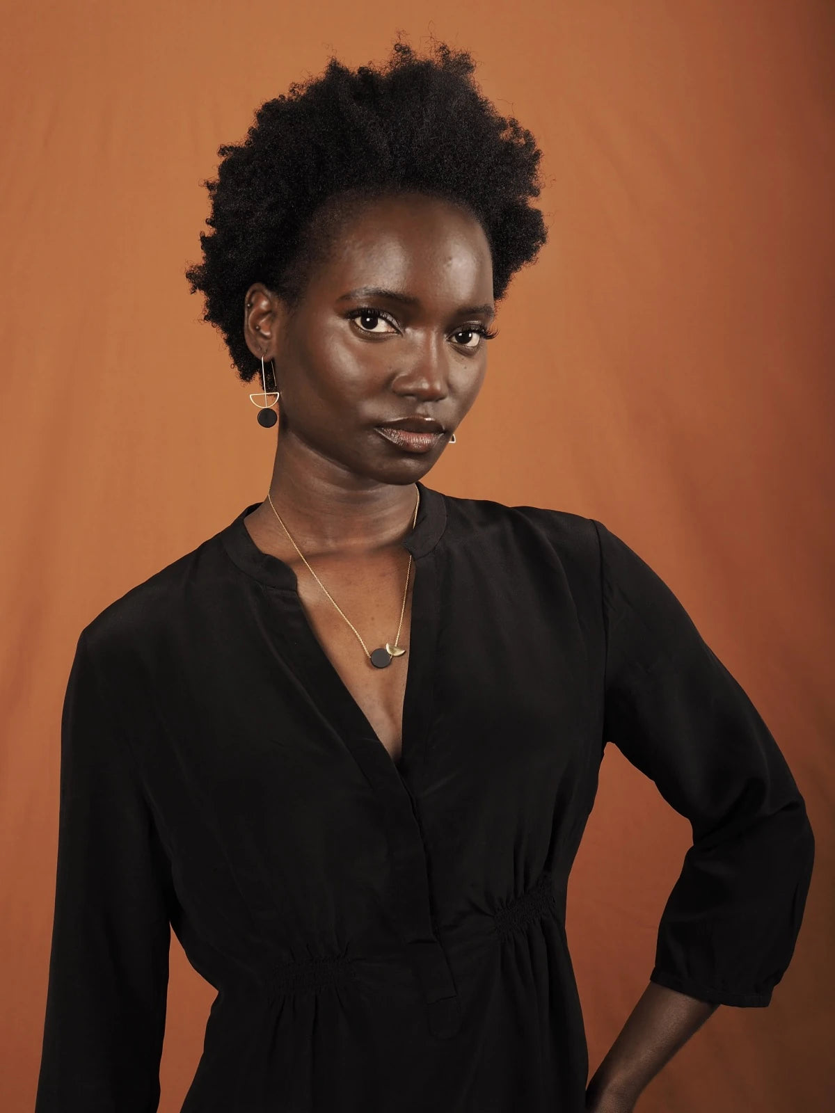 A woman with short curly black hair wearing a black long-sleeve V-neck dress wearing Nucleo Brass and Black Upside Down Arch Earrings by brass+bold, posing against a solid orange backdrop.