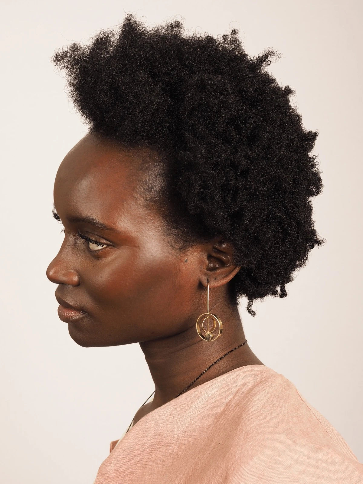 A woman in profile wearing a peach-pink top and Nucleo Brass Rings within Rings Earrings by brass+bold, showcasing her natural dark curly hair against a plain light beige background.