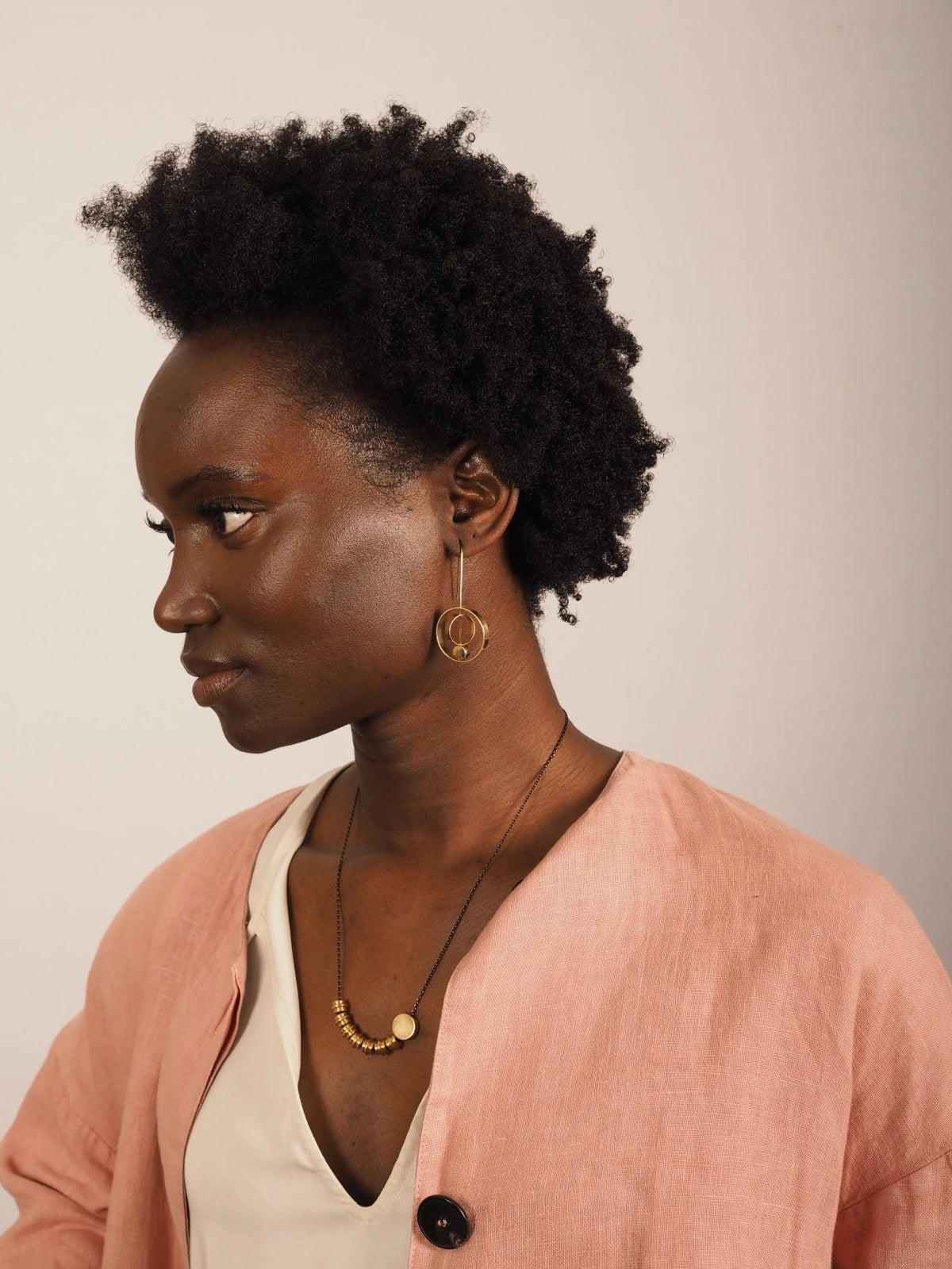 A woman with short, curly black hair is shown in profile wearing Nucleo Brass Rings within Rings Earrings by brass+bold, a delicate gold necklace, and a light pink cardigan over a cream top, set against a plain light beige background.