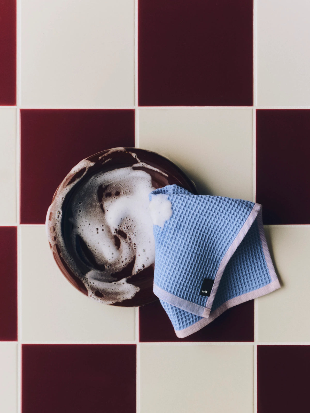 A blue waffle-weave HAY Canteen dishcloth with white trim is draped over a round brown dish covered in white foam, set against a dark red and cream checkered tile background.