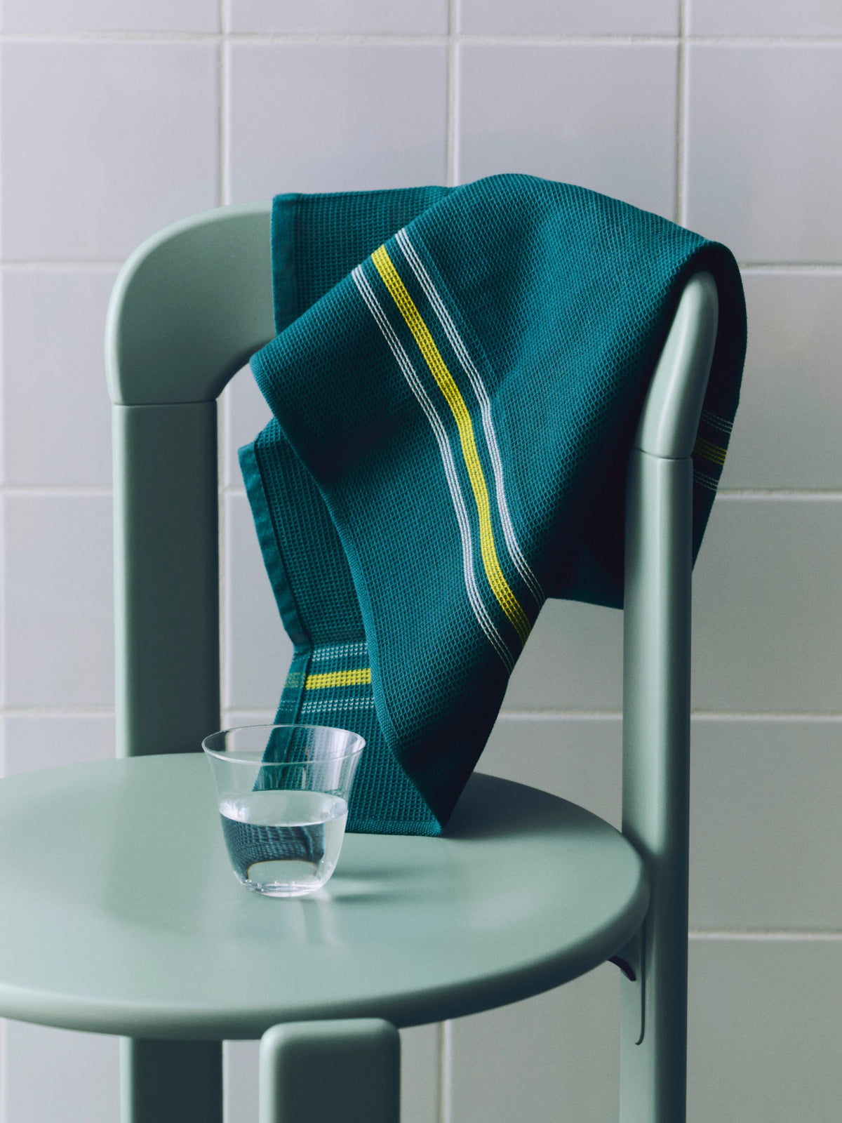 A dark teal HAY Canteen kitchen towel with yellow and white striped patterns draped over a matching sage green chair, with a small clear glass of water on the chair seat against a tiled background.