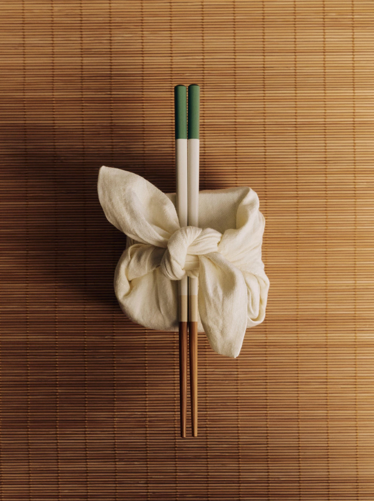 A pair of HAY Colour Sticks Chopsticks with green and white handles tied with a cream fabric bow, placed on a brown bamboo mat.