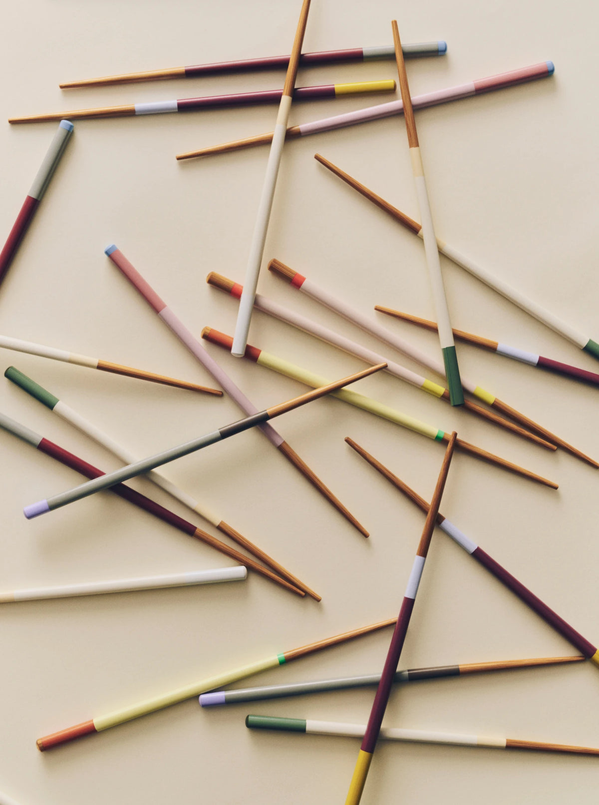 A scattered arrangement of wooden HAY Colour Sticks Chopsticks with painted handles in various colours, including brown, white, pink, yellow, green, and purple, on a light beige background.
