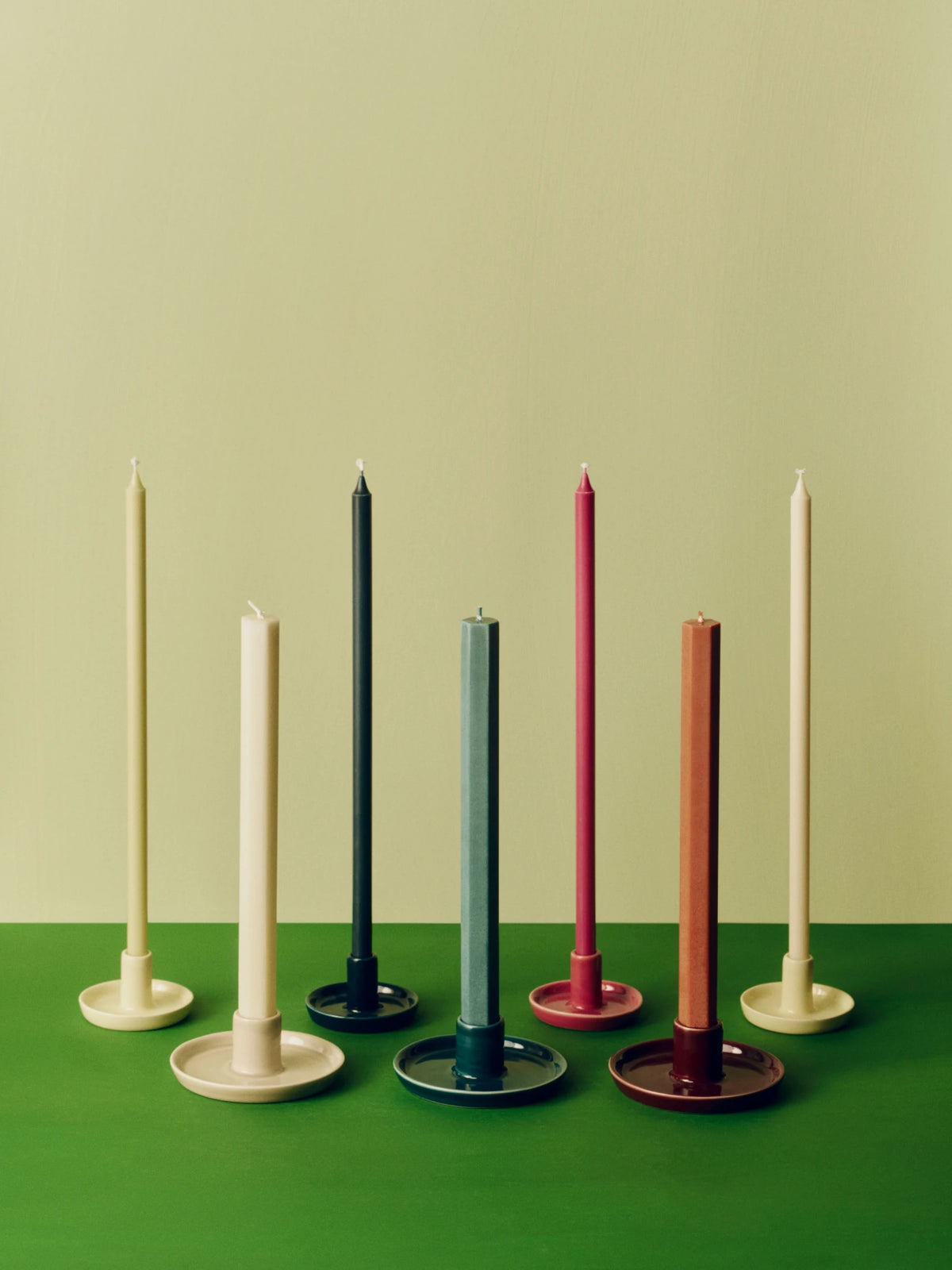 Six slim taper candles in different colors standing in matching HAY disc candle holders on a green surface against a pale green background.