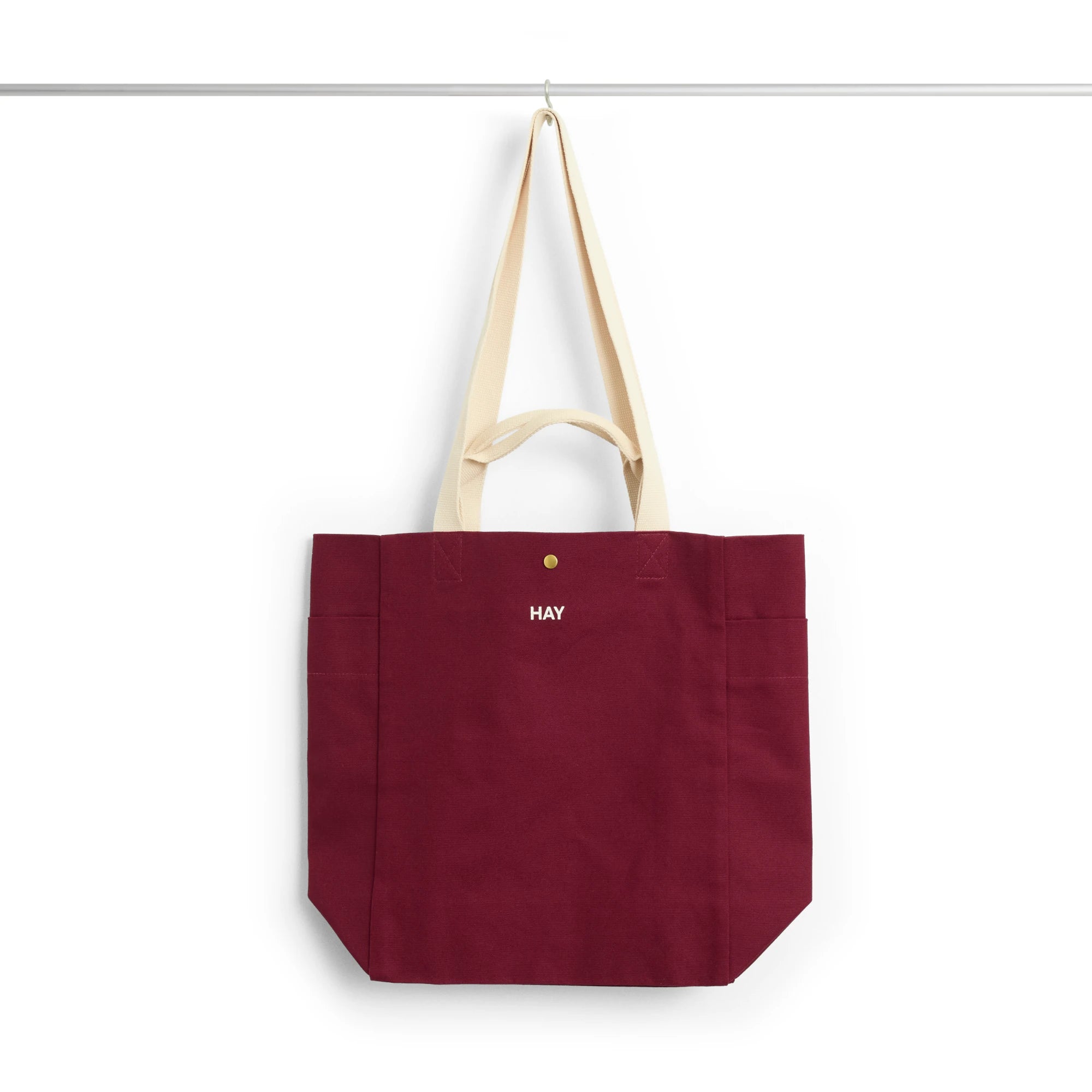 HAY burgundy canvas medium everyday tote bag with cream-colored shoulder straps and a small white 'HAY' logo on the front, hanging from a thin horizontal bar against a white background.