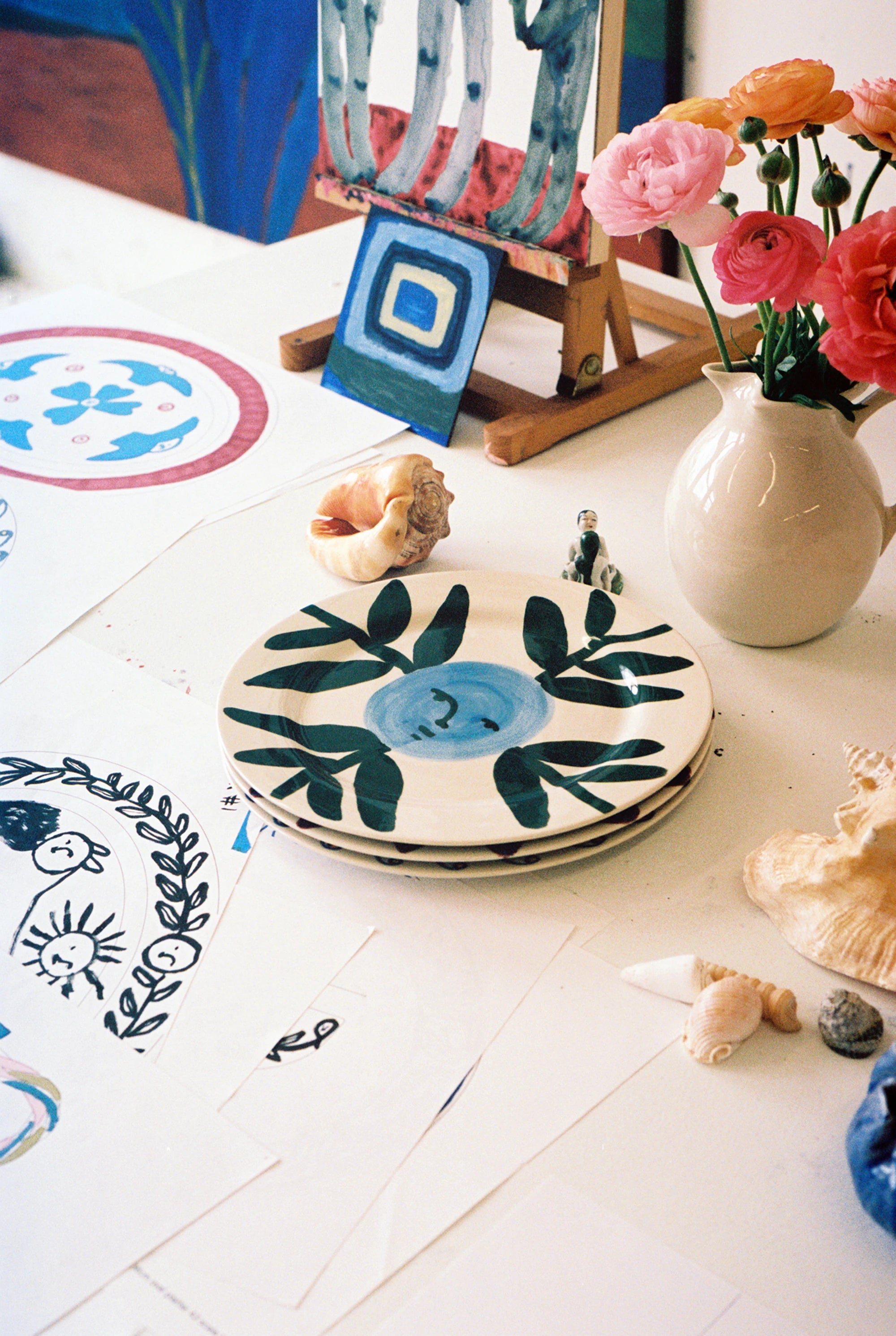 HAY x Emma Kohlmann - La Pittura Moon Face Vine plate stack on a busy studio work table beside drawing, a vase of flowers and an easel 