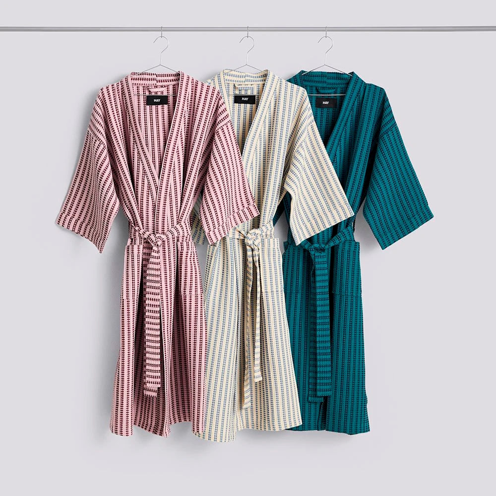 Three striped HAY waffle bathrobes hanging on hangers against a light gray wall: one soft pink, one blue and white, and one petrol blue.
