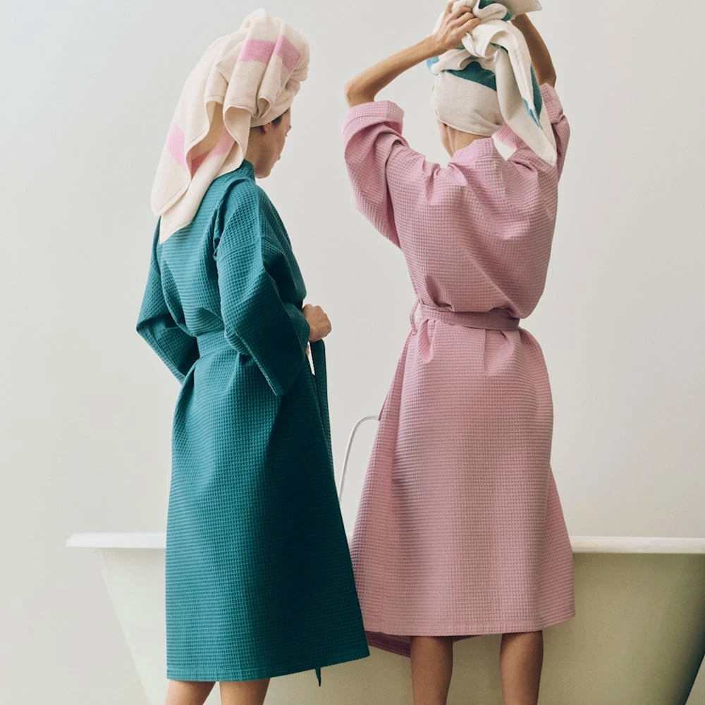 Two women wearing textured bathrobes stand in front of a white bathtub, seen from behind. One robe is teal green and the other is dusty pink, both with checkered waffle patterns.