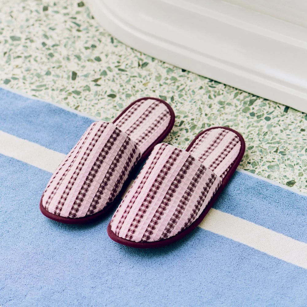 A pair of HAY striped waffle slippers in cool pink, placed on a blue rug with a green speckled tile floor in the background.