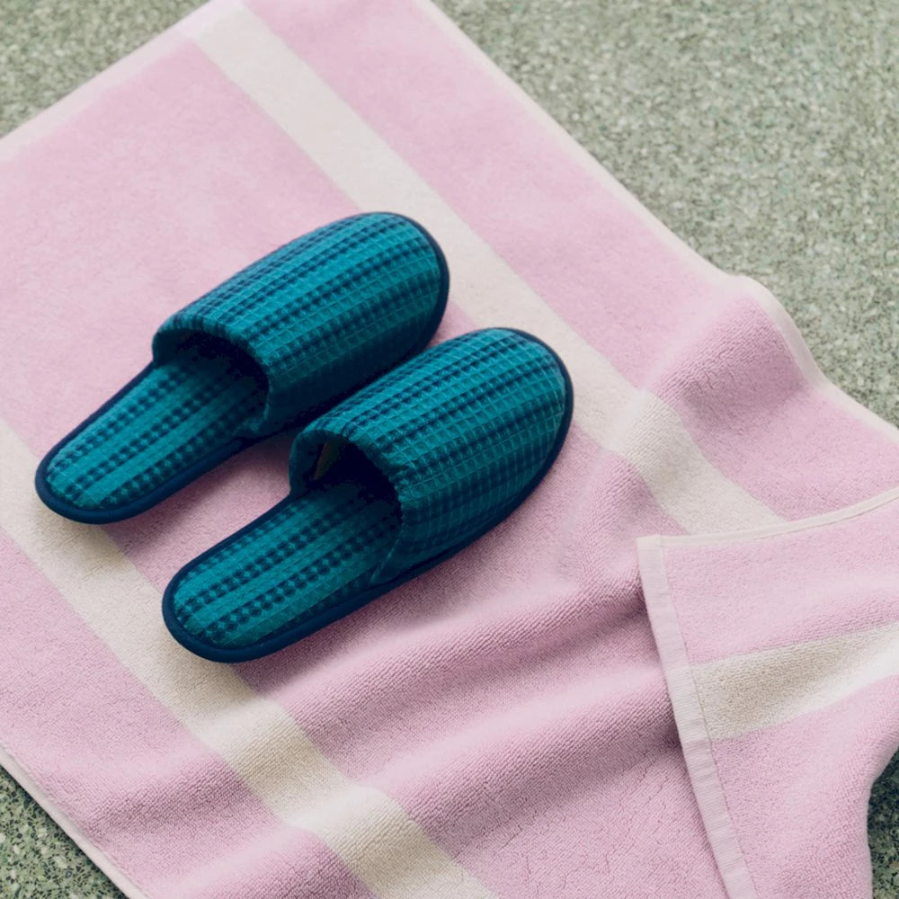 A pair of teal blue checkered slip-on slippers placed on a pink and white striped towel on a gray textured floor.
