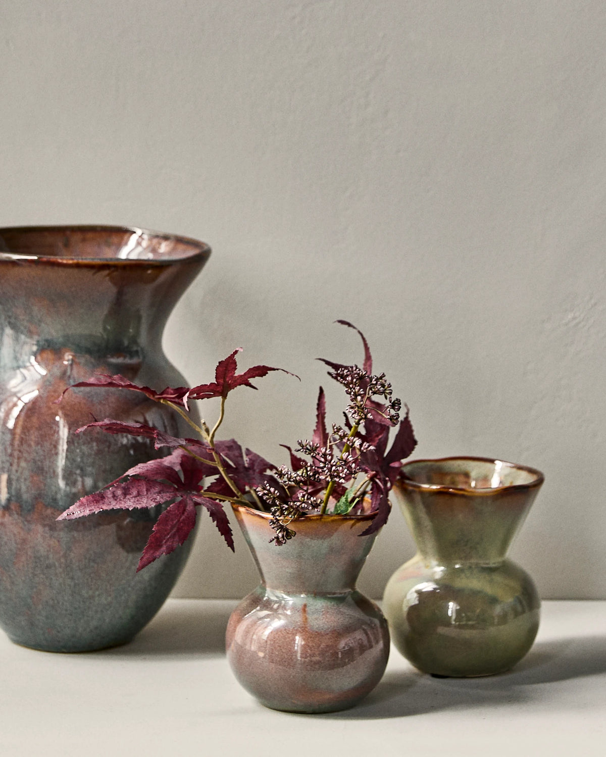 Three handmade ceramic vases with rustic glazes in earthy tones, arranged on a white surface against a neutral grey wall. The set includes a large vase on the left, a small vase in the center holding burgundy foliage, and a smaller House Doctor HDCorp Vase in Sea Green on the right.
