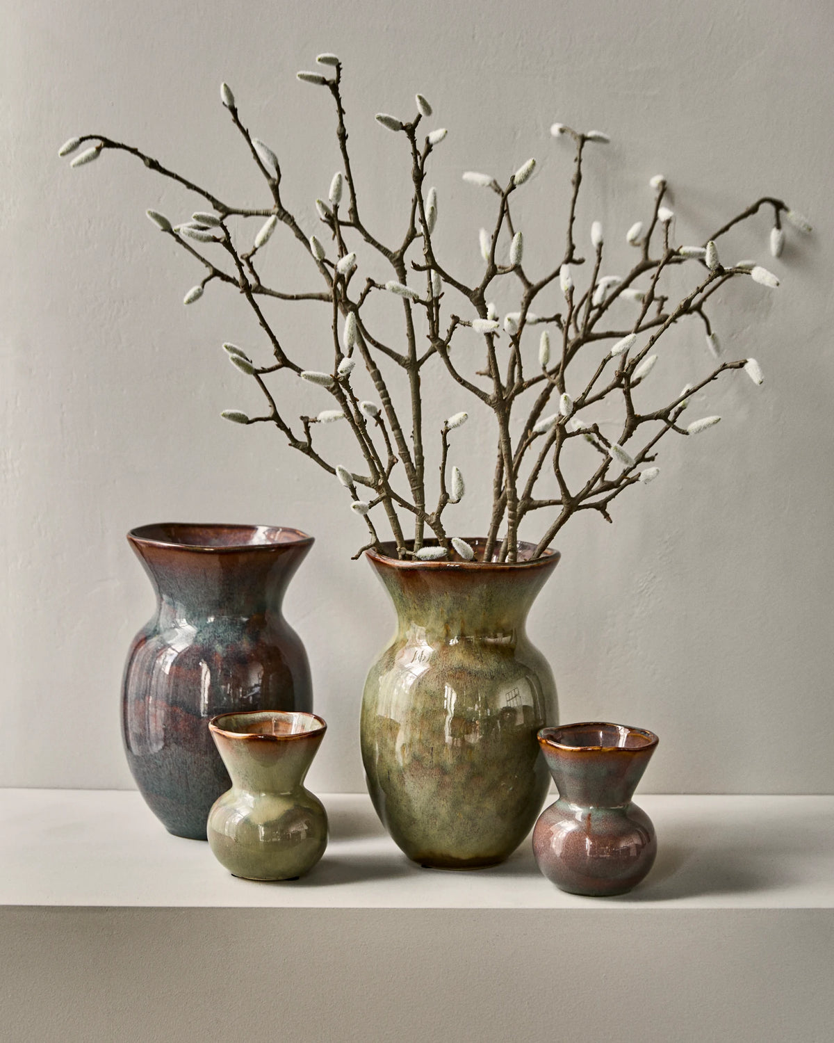 Four rustic glazed ceramic vases, including House Doctor HDCorp Vase in Sea Green in earthy tones arranged on a white shelf against a neutral grey wall, with a decorative branch arrangement in the tallest vase.