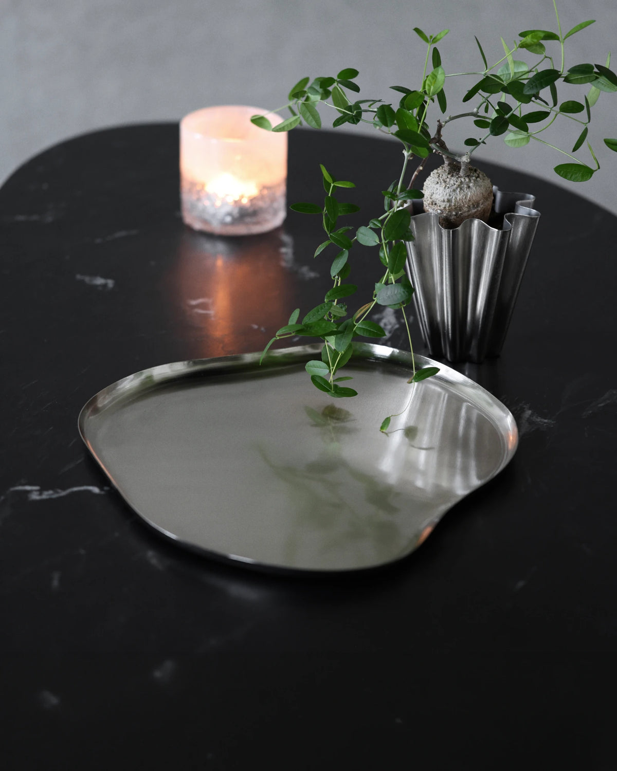 An organic-shaped House Doctor HDOsea Tray, Silver Finish with a polished reflective surface, styled on a dark marble tabletop next to a lit candle and a small potted green plant.