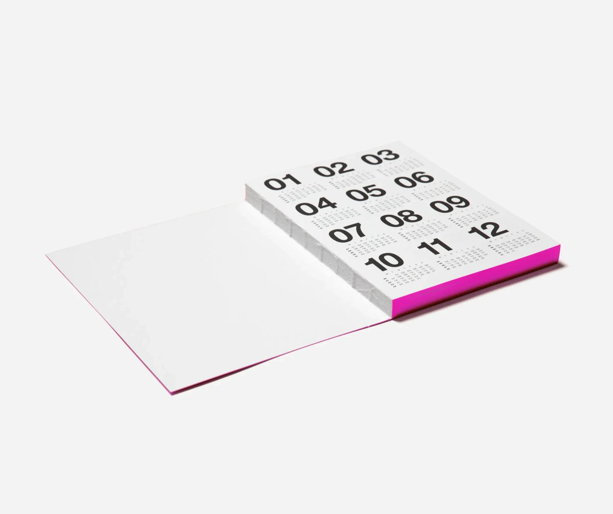 A minimalist open Marjolein Delhaas planner on a light grey background, showing a white interior spread with black numbers from 01 to 12 on the page and a bright pink edge.