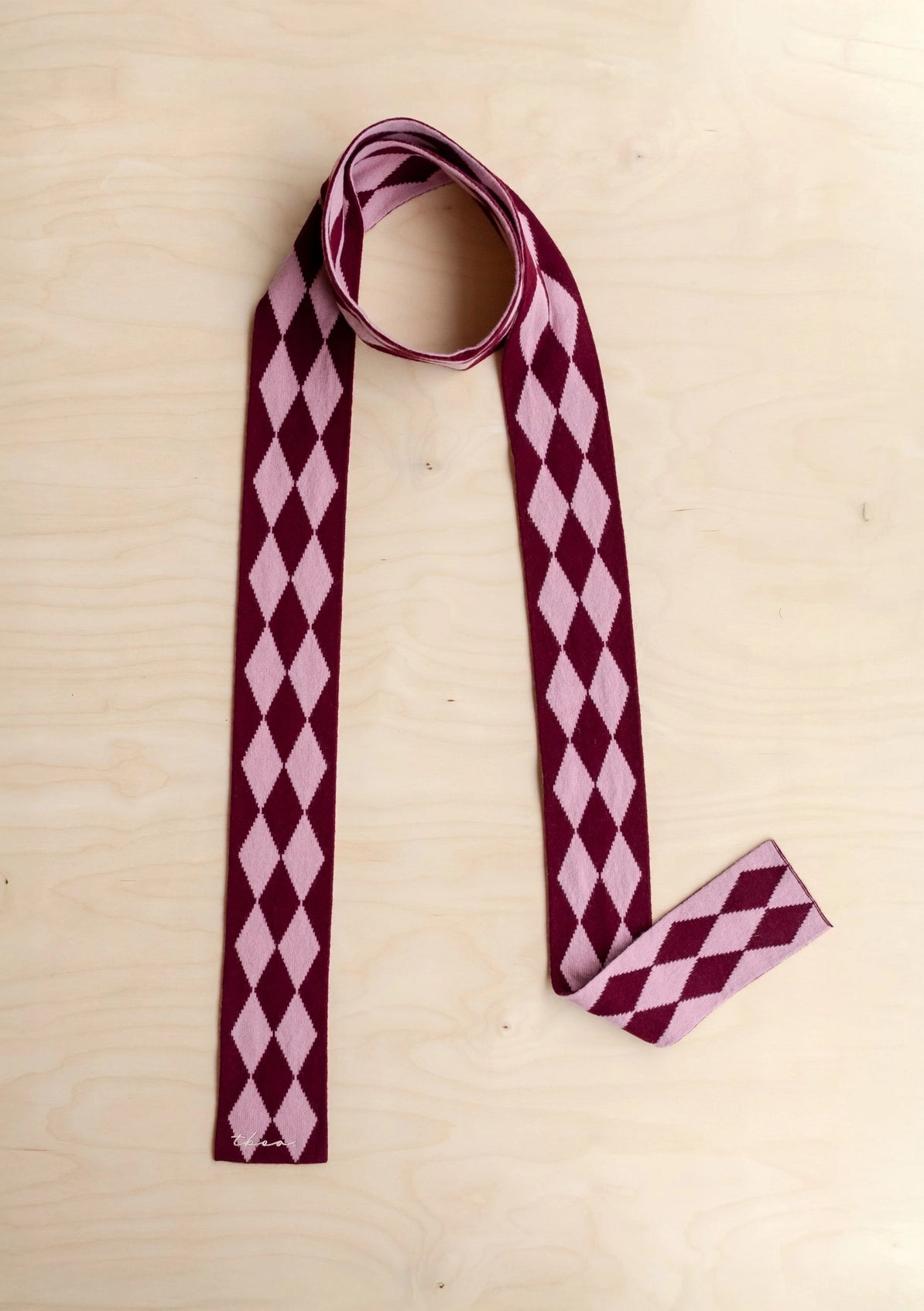 A long skinny scarfwith a classic harlequin diamond pattern in deep burgundy and light pink, laid flat on a light wood background.