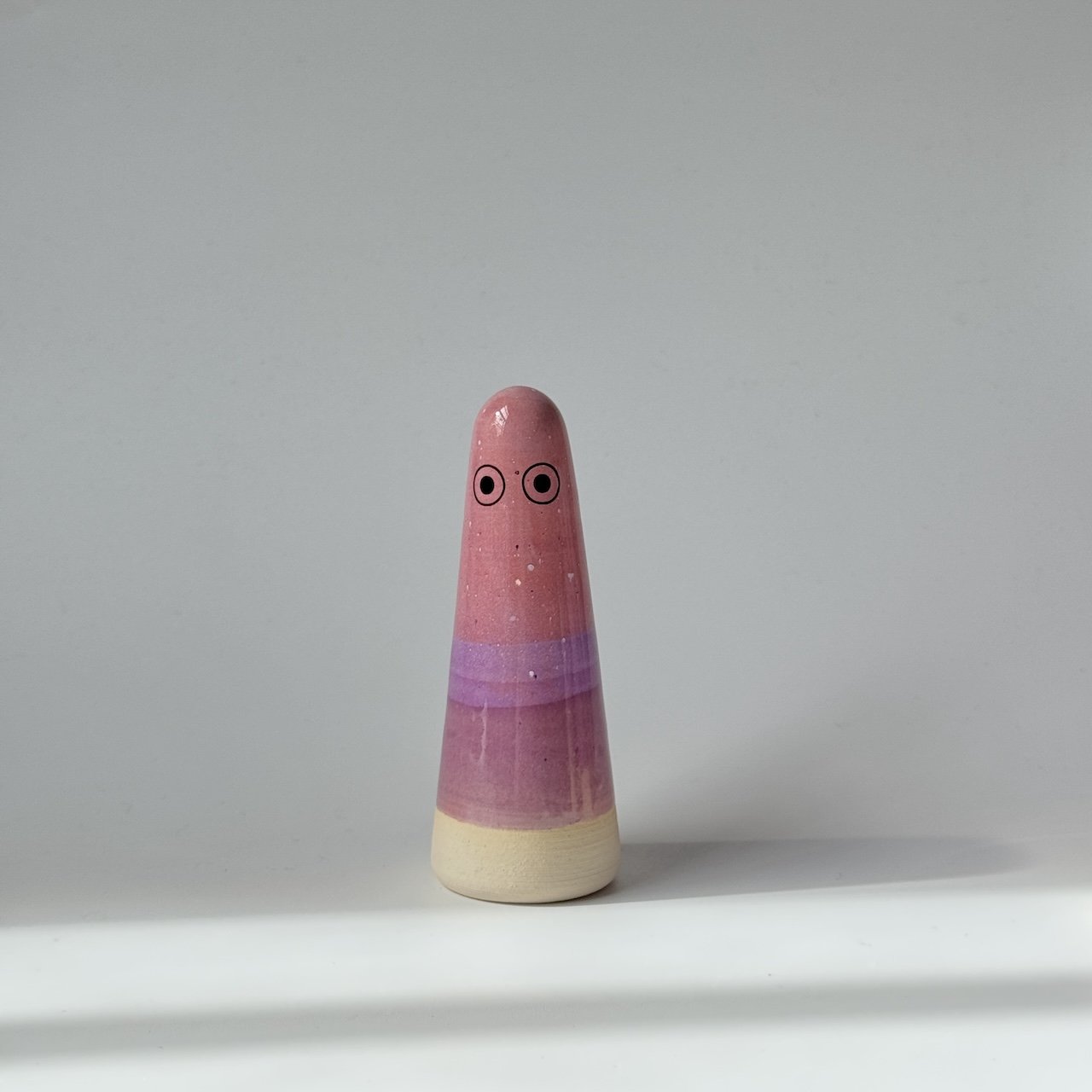 Ely Studio Arhoj ghost, featuring a pink and purple glazed body with a natural beige wooden base and simple painted eyes, set against a plain light grey background.