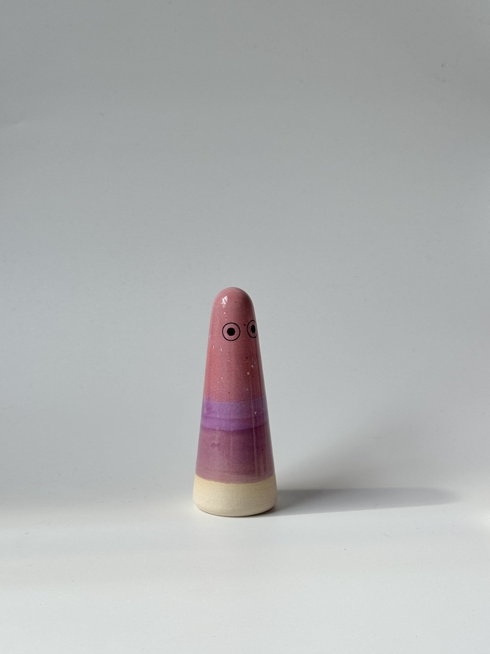 Ely Studio Arhoj ghost, featuring a pink and purple glazed body with a natural beige wooden base and simple painted eyes, set against a plain light grey background.